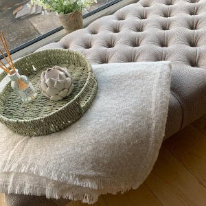 Pode incluir: Um cobertor cinza claro com borda franjada está drapeado sobre um otomano estofado. Uma bandeja de vime com uma vela decorativa e uma garrafa de difusor "Fresh Linen" estão sobre o otomano.