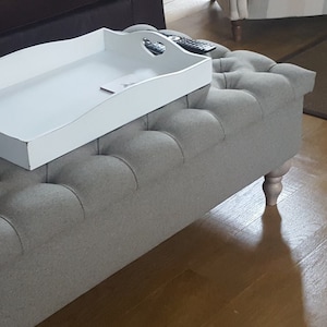 Puede incluir: Una bandeja de madera blanca con un asa de corte de corazón se encuentra en la parte superior de una otomana capitoné gris con patas de madera. La otomana está en un suelo de madera.