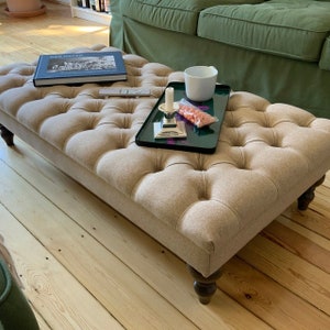 Op de afbeelding: Een beige getufte ottomane met vier houten poten. De ottomane is bedekt met een zwarte dienblad, een witte mok, een kleine kaars en een zakje oranje snoepjes. De ottomane staat op een houten vloer.