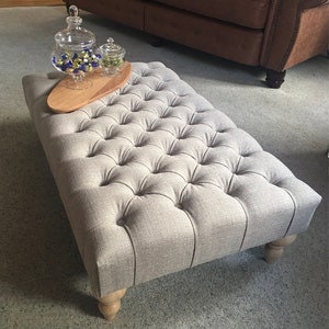 Può includere: Un pouf capitonné grigio con gambe in legno e un vassoio in legno in cima. Il pouf è rivestito in un tessuto grigio chiaro con un motivo sottile.