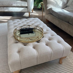 Pode incluir: Um otomano bege capitoné com uma bandeja de metal dourado no topo. A bandeja tem um fundo espelhado e um design decorativo ao redor da borda.