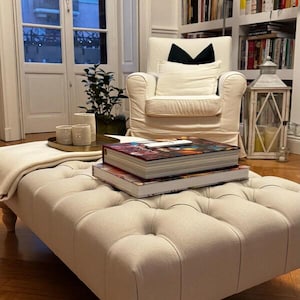 Pode incluir: Um pufe tufado bege numa sala de estar. Dois livros estão empilhados no pufe. Uma poltrona branca com uma almofada preta e uma lanterna decorativa estão no fundo. O pufe é retangular.