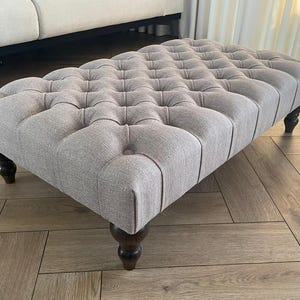 May include: A rectangular ottoman upholstered in light grey fabric with a button-tufted design. It features four dark brown, turned wooden legs. The ottoman is positioned on a wooden floor.