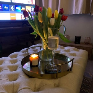 Peut inclure: Un plateau doré avec une bougie allumée, une coupe de champagne et un vase de tulipes colorées sur un pouf blanc.
