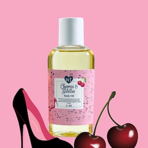 Op de afbeelding: Een heldere glazen fles body oil met een witte dop, met het label "Cherries & Stilettos". Het label is roze met kersen- en stiletto-afbeeldingen. Een zwarte en roze stiletto en twee kersen staan ook in de afbeelding. De fles bevat 59 ml olie.