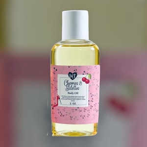 Op de afbeelding: Een heldere glazen fles body oil met een witte dop. Het roze etiket leest "Cherries & Stilettos" en heeft een kersen illustratie. De fles bevat 59 ml olie.