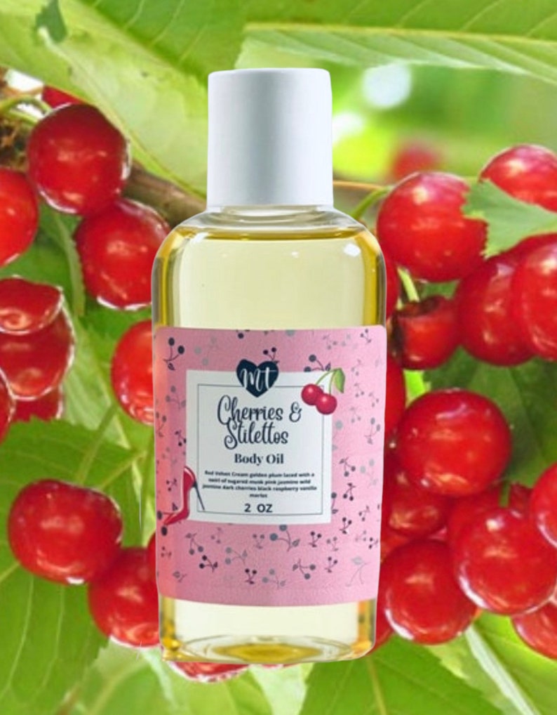 Op de afbeelding: Een heldere glazen fles body oil met een witte dop. Het roze etiket leest "Cherries & Stilettos Body Oil" met een kersenillustratie. De achtergrond toont rode kersen en groene bladeren. De fles bevat 59 ml olie.