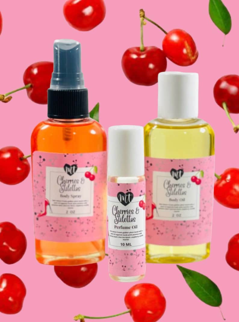 Op de afbeelding: Drie flessen geurproducten met roze etiketten en de tekst "Cherries & Stilettos". De flessen zijn gevuld met oranje en gele vloeistoffen. Rode kersen zijn verspreid rond de flessen tegen een roze achtergrond. De body spray fles is 59 ml.