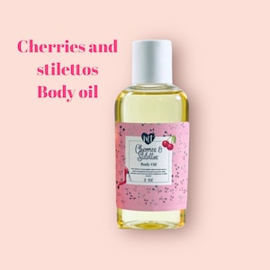 Op de afbeelding: Een heldere glazen fles body oil met een witte dop. Het etiket is roze met de woorden "Cherries and Stilettos Body Oil" en een kersenillustratie. De fles bevat 59 ml olie.