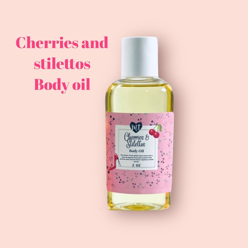Op de afbeelding: Een heldere glazen fles body oil met een witte dop. Het etiket is roze met de woorden "Cherries and Stilettos Body Oil" en een kersenillustratie. De fles bevat 59 ml olie.