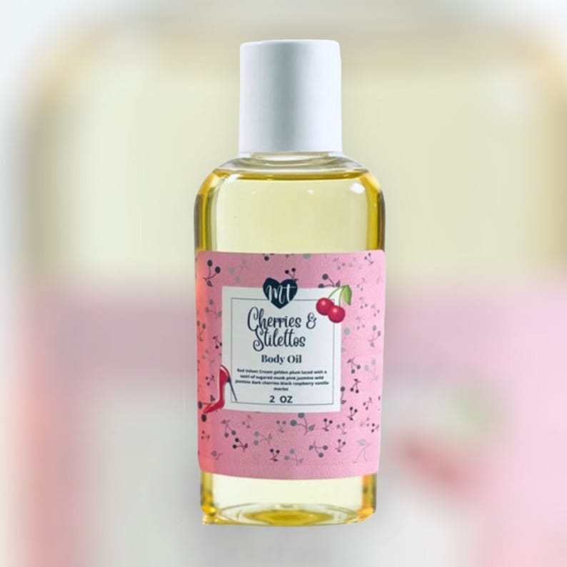 Op de afbeelding: Een heldere glazen fles gevuld met gele lichaamsolie. Het roze etiket bevat de woorden "Cherries & Stilettos" en een kersenillustratie. De fles heeft een witte dop en bevat 60 ml product.