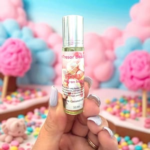 Op de afbeelding: Een heldere glazen roll-on fles met een zilveren dop, vastgehouden door een hand met lichtpaarse nagellak. De fles heeft een label met de tekst "Tresor Beaut" en "Roll-on body". De achtergrond is een kleurrijke, vage scène met roze en blauwe elementen.