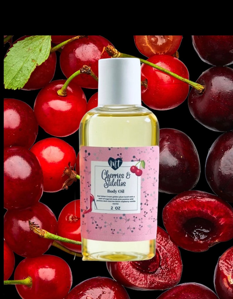 Op de afbeelding: Een heldere glazen fles body oil met een witte dop, met het label "Cherries & Stilettos". Het label is roze met een kersenillustratie. De achtergrond is een zwart oppervlak bedekt met verse rode kersen.