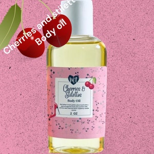 Op de afbeelding: Een kleine fles heldere body oil met een witte dop. Het etiket toont "Cherries & Stilettos Body Oil" met een kersen- en stiletto-afbeelding. De fles is 59 ml en staat tegen een roze achtergrond.