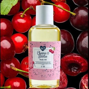 Op de afbeelding: Een heldere glazen fles body oil met een witte dop, met het label "Cherries & Stilettos". Het label is roze met een kersenillustratie. De achtergrond is een zwart oppervlak bedekt met verse rode kersen.