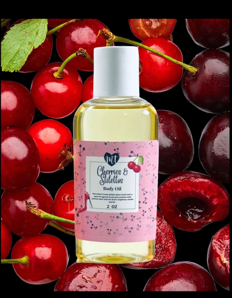 Op de afbeelding: Een heldere glazen fles body oil met een witte dop, met het label "Cherries & Stilettos". Het label is roze met een kersenillustratie. De achtergrond is een zwart oppervlak bedekt met verse rode kersen.