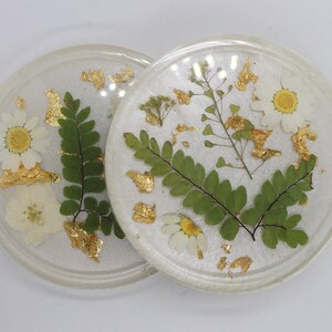 Puede incluir: Dos posavasos redondos de resina transparente con flores secas y detalles de pan de oro. Cada posavasos presenta margaritas blancas, helechos verdes y otros elementos botánicos delicados. Los posavasos tienen un acabado brillante.