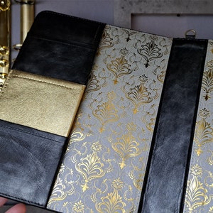 May include: A black leather-bound journal with a gold-embossed design. The journal has a gold-colored pocket on the front cover and a black ribbon bookmark.