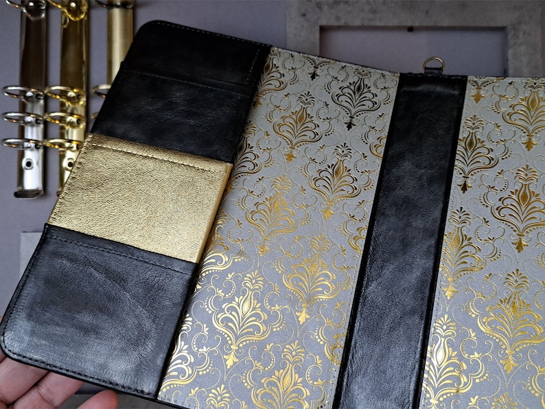 May include: A black leather-bound journal with a gold-embossed design. The journal has a gold-colored pocket on the front cover and a black ribbon bookmark.