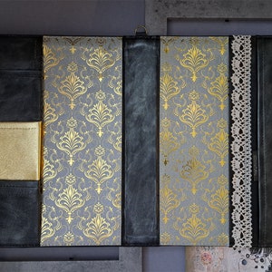 May include: A black leather journal with a gold patterned cover and a lace trim. The journal has a gold pocket on the front cover.