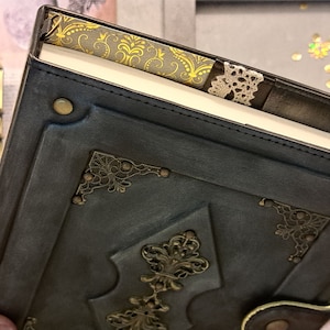 May include: A dark blue leather-bound journal with decorative gold metal accents and a lace bookmark. The journal has a vintage aesthetic.