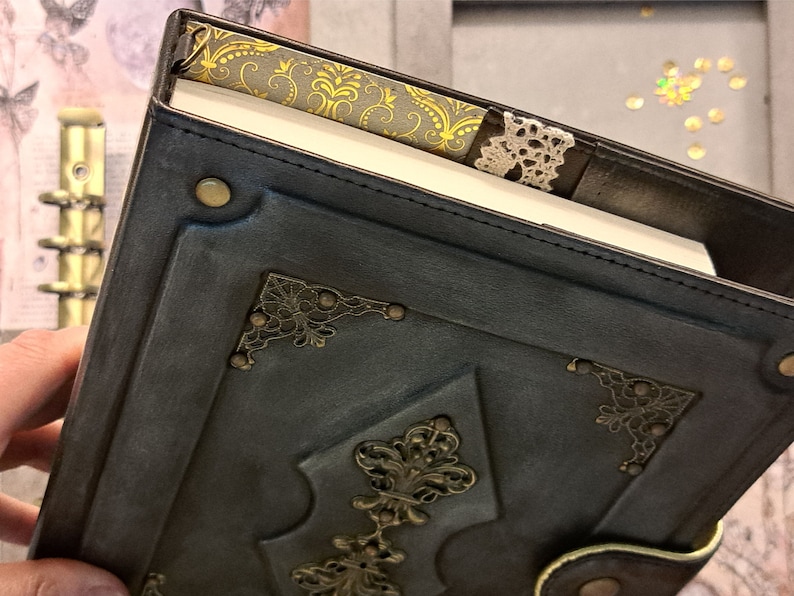 May include: A dark blue leather-bound journal with decorative gold metal accents and a lace bookmark. The journal has a vintage aesthetic.