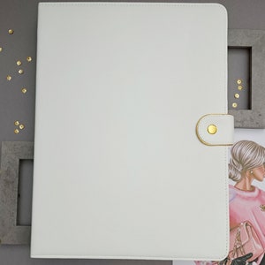 Può includere: Cartella in similpelle bianca con chiusura a scatto dorata. La cartella è accanto a un'illustrazione incorniciata di una donna con un maglione rosa e guanti neri. Paillettes dorate sparse.