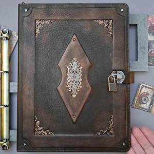 May include: A dark brown leather journal with a decorative metal clasp and lock. The cover features a central diamond-shaped design with ornate silver detailing and bronze corner accents. Includes a gold binder and decorative cards.