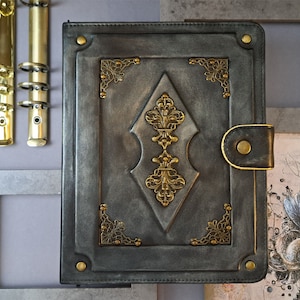 May include: A black leather-bound journal with ornate gold accents and a decorative diamond-shaped design. The journal has a flap closure with a metal button.