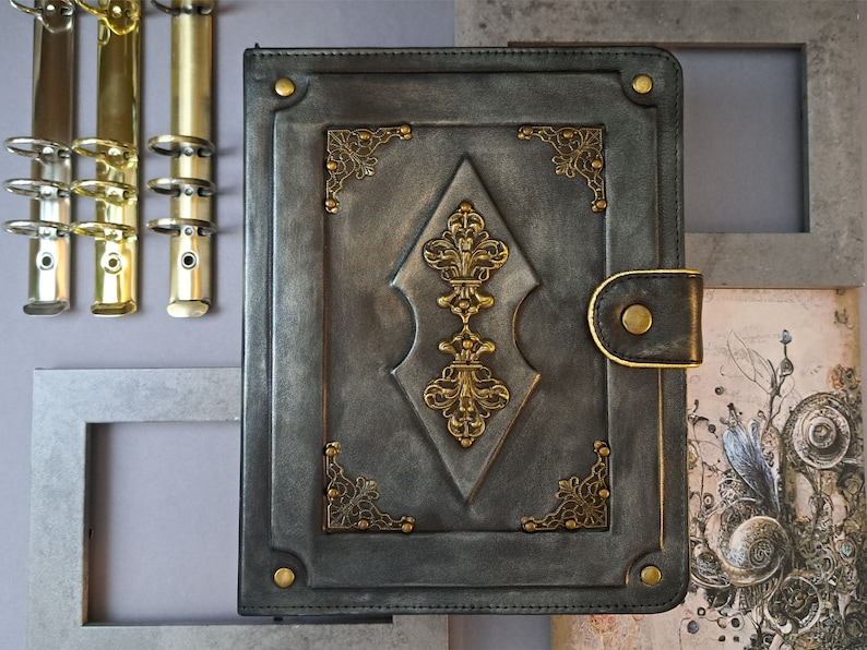 May include: A black leather-bound journal with ornate gold accents and a decorative diamond-shaped design. The journal has a flap closure with a metal button.