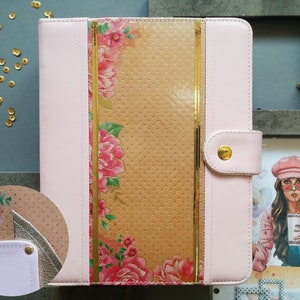Può includere: Un'agenda in pelle rosa con dettagli dorati e un design floreale. L'agenda ha una cerniera dorata e una chiusura dorata.