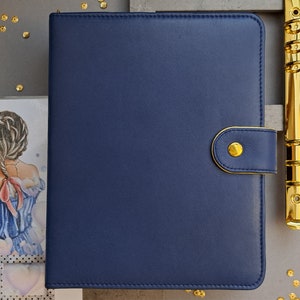 Może przedstawiać: Planer w ciemnoniebieskiej skórzanej oprawie z pozłacanym metalowym binderem i złotym guzikiem. Planer ma klapkę zakrywającą pierścienie.