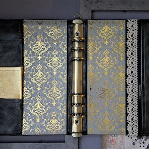 May include: A black leather bound journal with a gold and gray patterned cover. The journal has a brass ring binder and a lace trim.