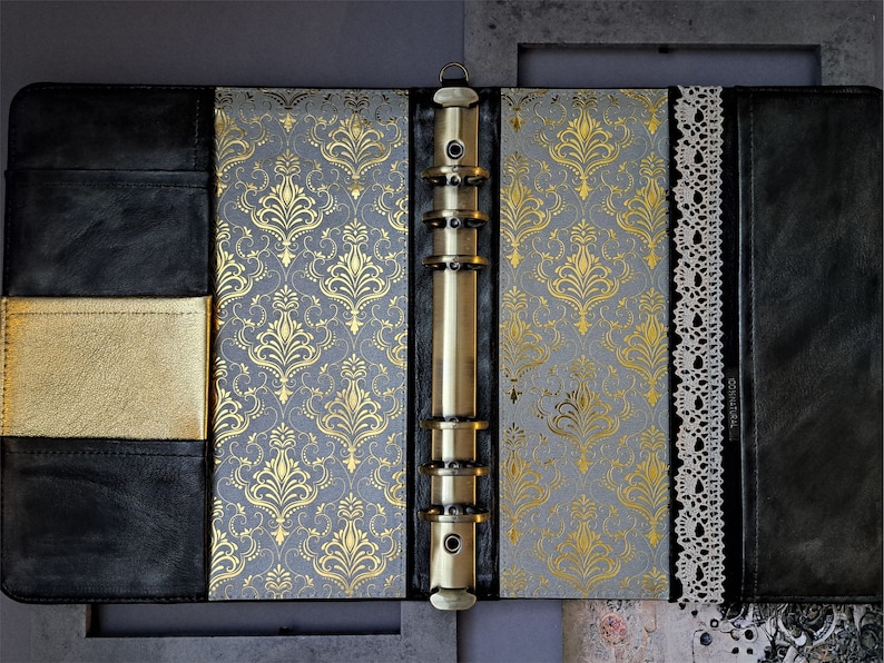 May include: A black leather bound journal with a gold and gray patterned cover. The journal has a brass ring binder and a lace trim.