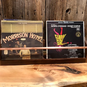 May include: A dark wood record stand with a copper bar across the front. Two vinyl records are displayed on the stand. The record on the left is "The Doors - Morrison Hotel". The record on the right is "Funny Girl" starring Barbra Streisand and Sydney Chaplin.