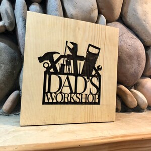 May include: A wooden sign with the text "DAD'S WORKSHOP" in black lettering. The sign features a silhouette of a toolbox with various tools, including a hammer, saw, and wrench.