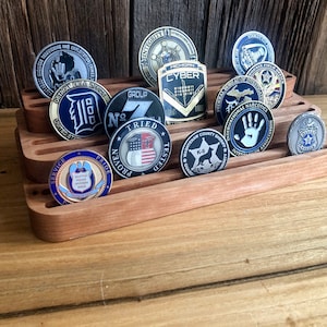K&ouml;nnte beinhalten: Ein dreistufiger Holzst&auml;nder zeigt eine Sammlung von Challenge-M&uuml;nzen. Die M&uuml;nzen zeigen verschiedene Designs, darunter Motive aus den Bereichen Strafverfolgung, Milit&auml;r und Sport. Die M&uuml;nzen sind aus Metall und haben verschiedene Oberfl&auml;chen, darunter Gold, Silber und Bronze.