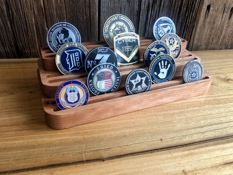 K&ouml;nnte beinhalten: Ein dreistufiger Holzst&auml;nder zeigt eine Sammlung von Challenge-M&uuml;nzen. Die M&uuml;nzen zeigen verschiedene Designs, darunter Motive aus den Bereichen Strafverfolgung, Milit&auml;r und Sport. Die M&uuml;nzen sind aus Metall und haben verschiedene Oberfl&auml;chen, darunter Gold, Silber und Bronze.