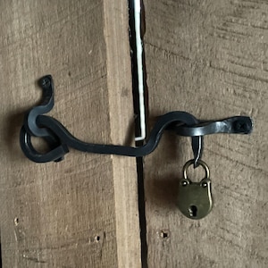 Puede incluir: Un pestillo de puerta de metal negro con un asa de bucle decorativo y un pequeño candado de latón. El pestillo está unido a una puerta de madera.