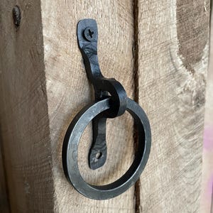 Op de afbeelding: Zwarte metalen ringtrekhengsel bevestigd aan een houten deur. De hendel is een enkele ring met een gebogen metalen arm die aan de deur is bevestigd.