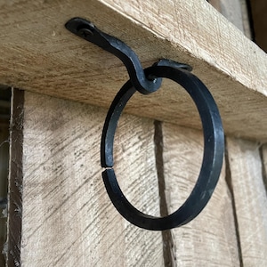 Könnte beinhalten: Schwarzer Metallringhaken, der mit einer Schraube an einem Holzregal befestigt ist. Der Haken ist zum Aufhängen von Gegenständen gedacht.