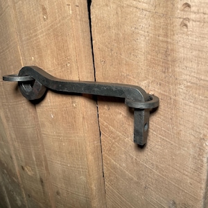 Könnte beinhalten: Ein schwarzer Türriegel aus Metall mit einem gebogenen Griff und einem quadratischen Sockel. Der Riegel ist an einer Holztür befestigt.