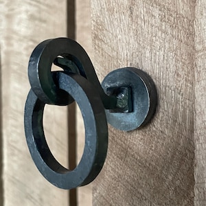 Może przedstawiać: Kuty żelazny uchwyt do drzwi z okrągłym uchwytem i okrągłą podstawą. Uchwyt jest połączony z podstawą za pomocą kwadratowego elementu. Okucia są zamontowane na drewnianych drzwiach.