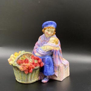 以下が含まれることがあります： 青いベレー帽と紫色のショールを着た女性が眠っている赤ちゃんを抱いている磁器製の置物。彼女は、カラフルな花が入ったバスケットの隣にあるベンチに座っています。