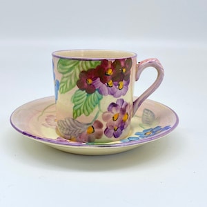 Puede incluir: Un juego de taza y platillo de té vintage con flores. La taza y el platillo son blancos con un patrón floral en tonos de morado, azul, verde y amarillo. La taza tiene un asa y el platillo tiene un borde festoneado.