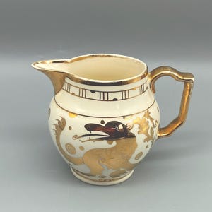 May include: Cream-colored ceramic pitcher with gold accents. The pitcher features a gold handle and spout, with a decorative gold dragon design. The rim and handle are also trimmed in gold. The pitcher has a bulbous shape.