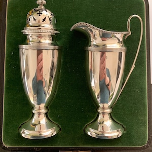 Puede incluir: Un juego de dos azucareros y jarras de crema de plata en una caja forrada de terciopelo verde. Los recipientes tienen forma de urnas con un diseño decorativo en la tapa del azucarero. La caja está etiquetada con "Regent Silver Ltd. London."
