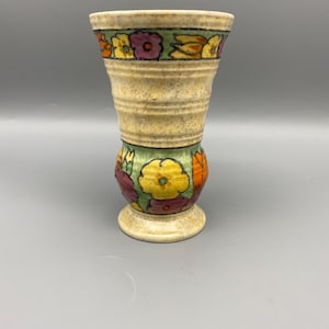 Op de afbeelding: Een vintage keramische vaas met een beige, getextureerd lichaam en een uitlopende bovenkant. De vaas is versierd met kleurrijke bloempatronen in geel, paars en oranje, geplaatst tegen groene banden. De basis heeft een bijpassende beige kleur.