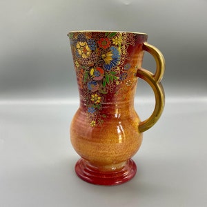 Puede incluir: Una jarra de cerámica roja y naranja con un diseño floral y detalles dorados. La jarra tiene dos asas y un borde acampanado.