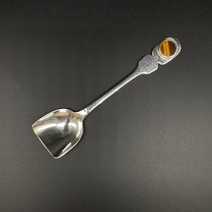May include: A silver spoon with a rounded, scoop-like bowl and a decorative handle. The handle is adorned with a round, brown tiger's eye gemstone.
